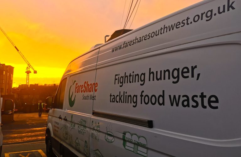 Close up of a FareShare South West-branded white van with a yellow and orange sunrise in the background and a crane silhouetted in the distance